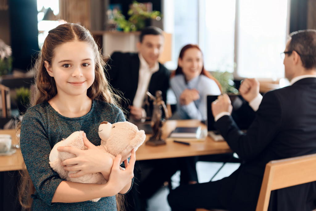 Young girl smiling while the parents are discussing custody with a lawyer in the background.